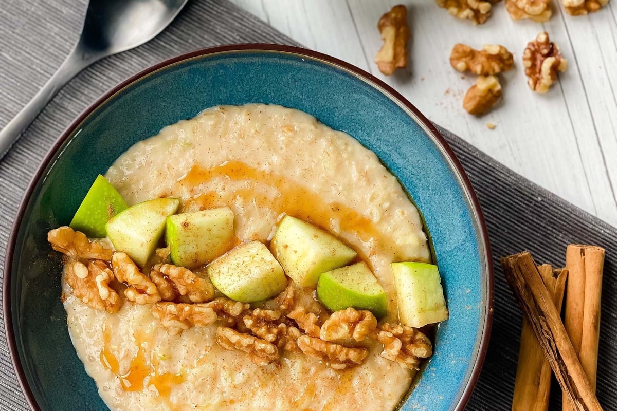 Bowl of porridge topped with walnuts, apple chunks, honey, and cinnamon sticks nearby