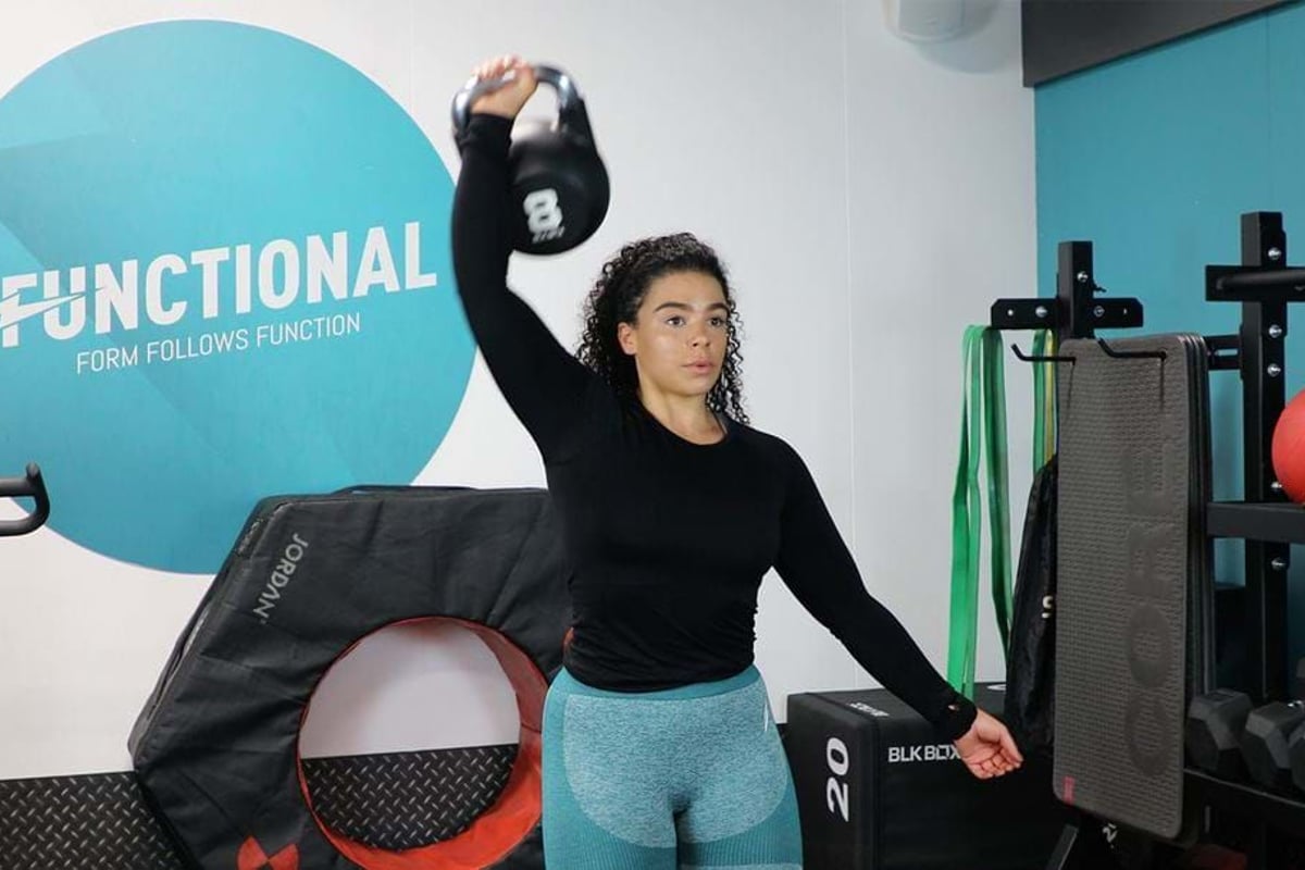 Woman lifting a kettlebell overhead in a gym with fitness equipment around her.