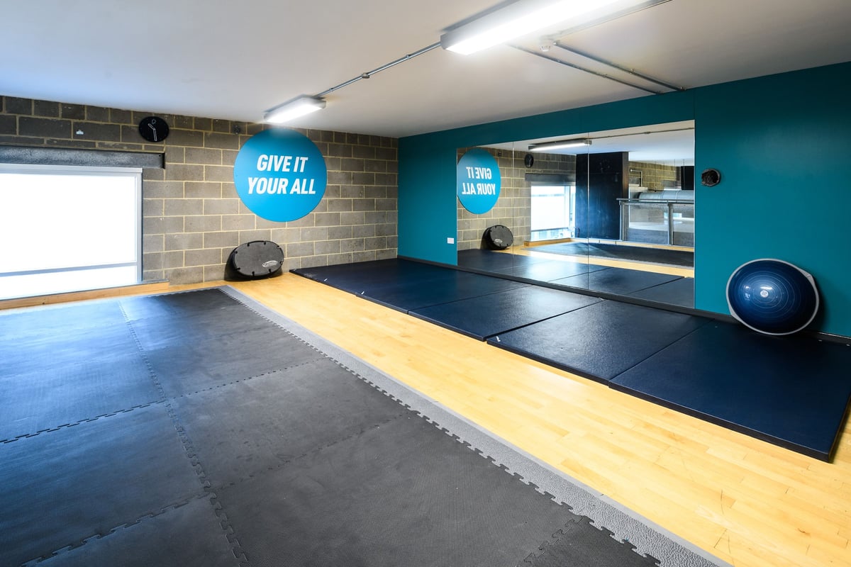 PureGym London Borough: Rubberized gym floor, mirrored walls, weightlifting platforms, stability balls, teal accents.