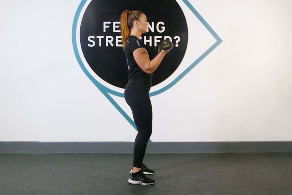 Woman in black workout clothes lifting dumbbells in front of a wall sign.