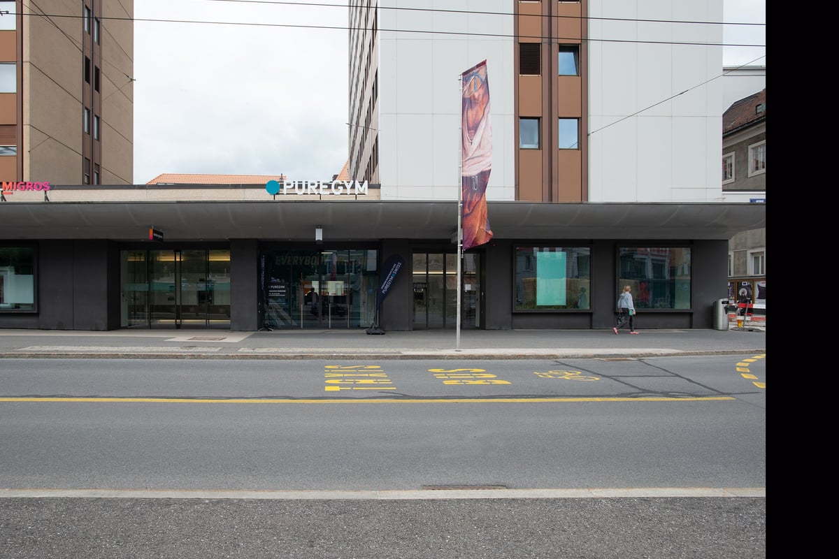 PureGym La Chaux-de-fonds: Small gym entrance with large windows, flags, and road markings outside on the street.