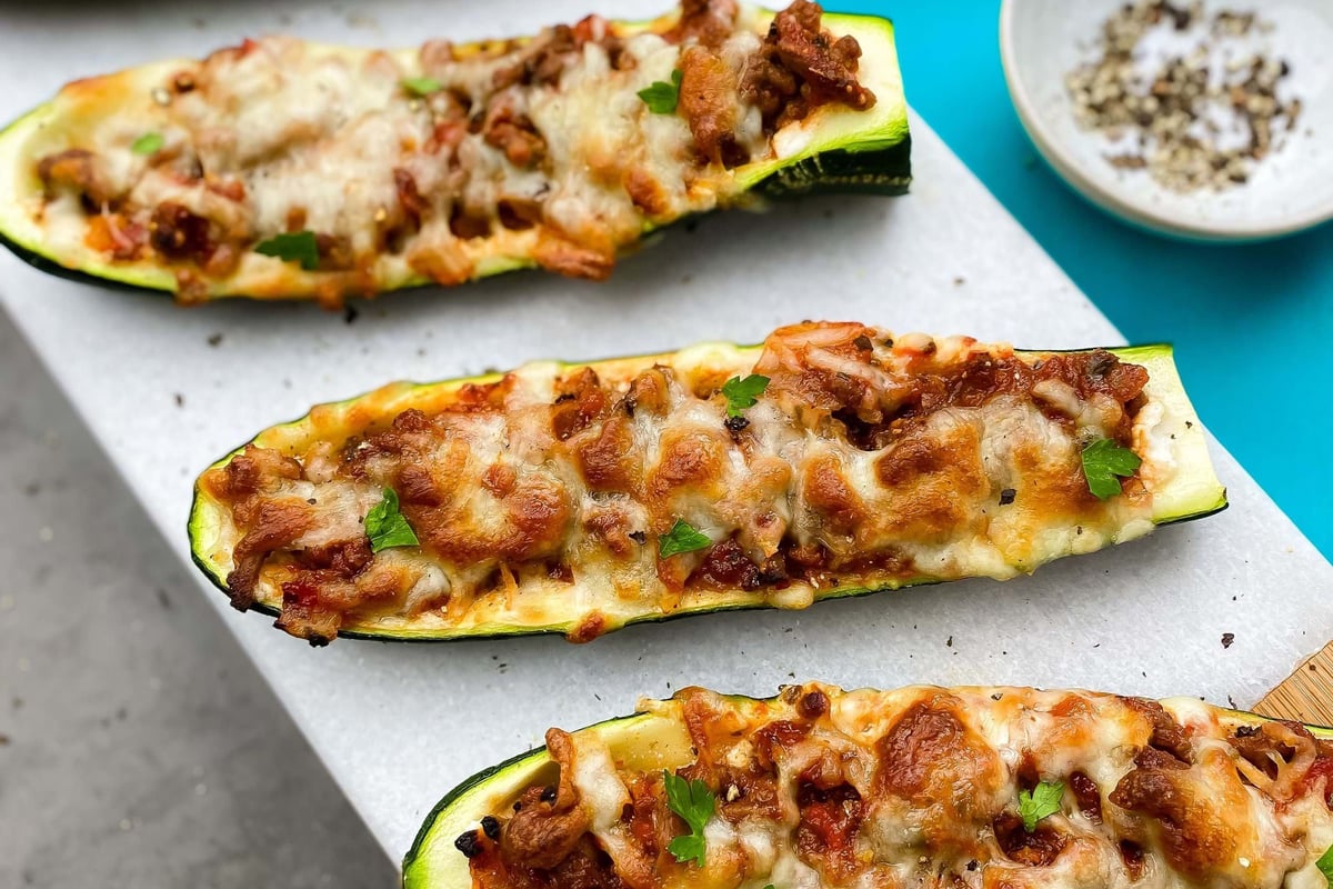 Four zucchini boats filled with meat and melted cheese on a white board with a bowl of sal