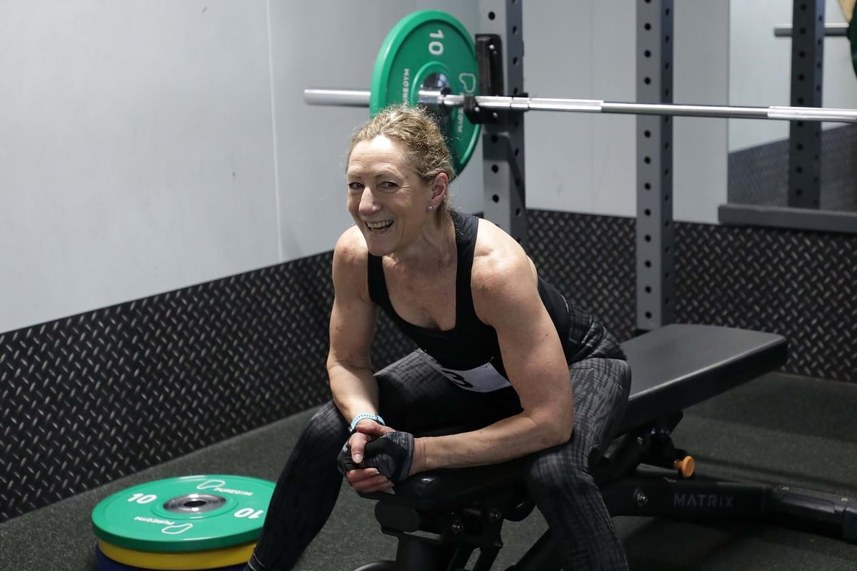 Woman in gym clothes sitting on bench with weights and barbell behind her smiling