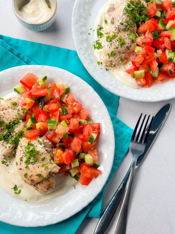 Grilled chicken with creamy sauce and tomato cucumber salad on white plates with cutlery.