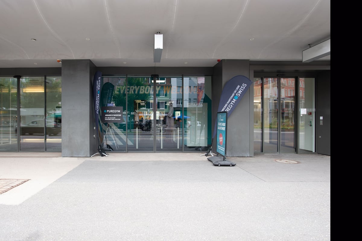 PureGym La Chaux-de-fonds: Empty gym entrance area with glass doors, flags, and branded signage visible.