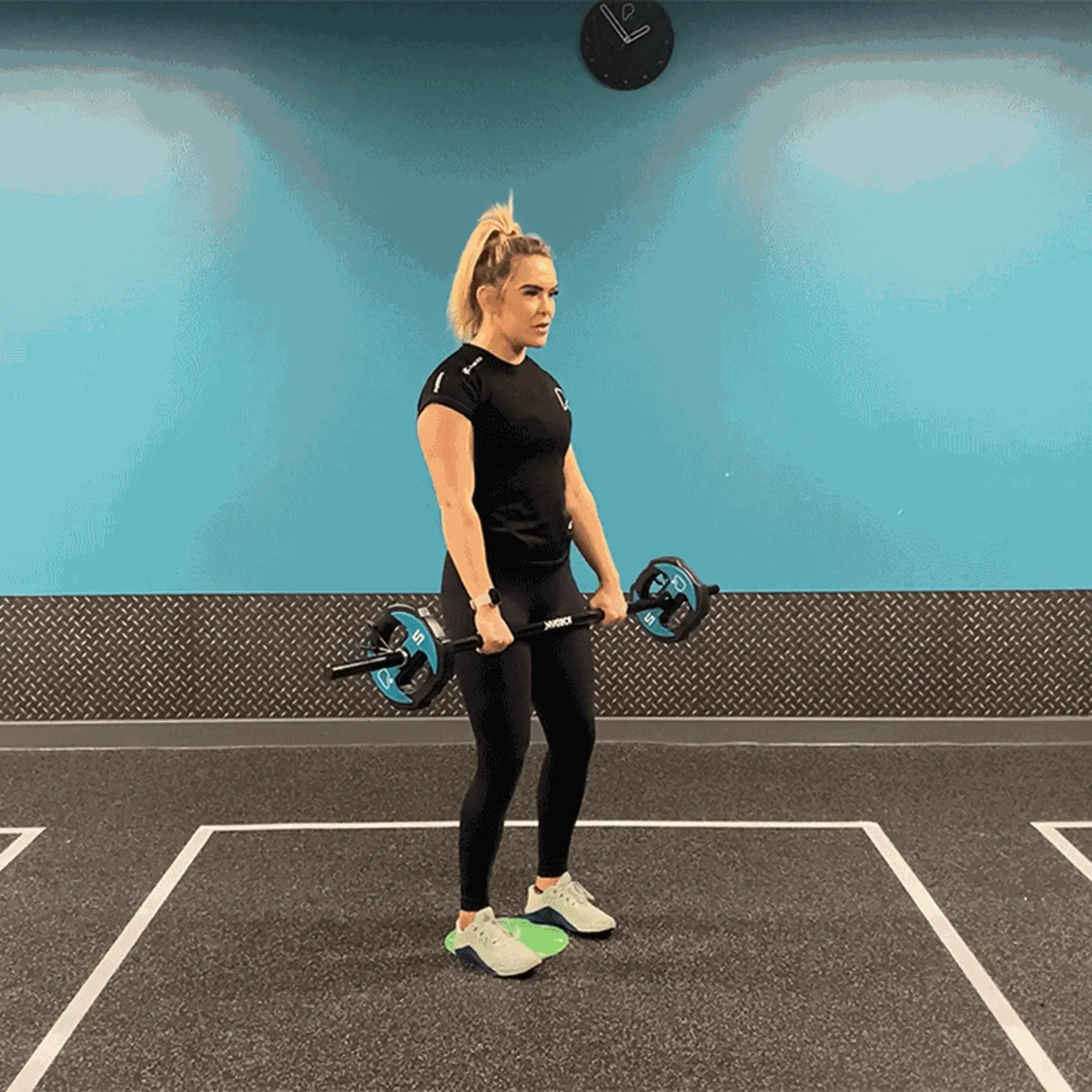 Woman in black sportswear lifting a barbell in a gym with blue walls and clock