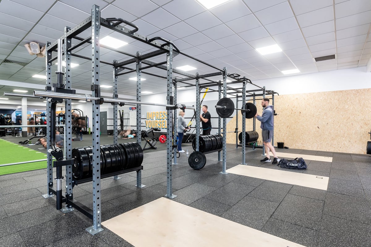 PureGym Woking: Squat racks, weight plates, rubberized turf area, and mirrored wall inside a gym studio.