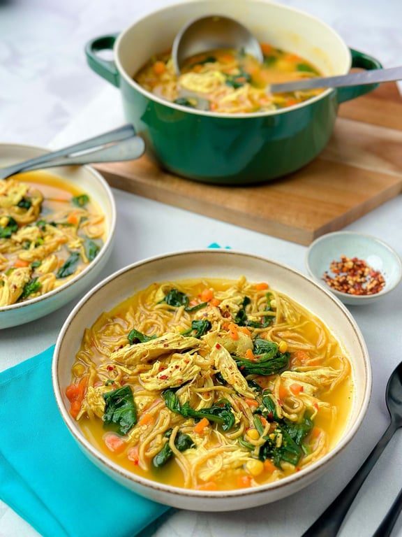 Bowls of chicken noodle soup with spinach and diced carrots on a tablecloth.