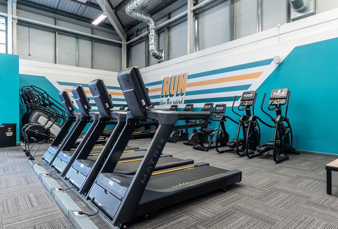 PureGym Kettering: Rows of treadmills, exercise bikes, and a vibrant mural lining the walls of a gym studio.