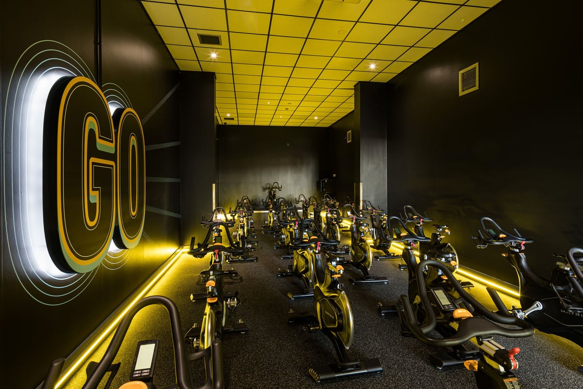 PureGym Dudley Tipton: Rows of stationary cycles in a dimly lit studio with yellow ceiling and neon 'Go' signage.