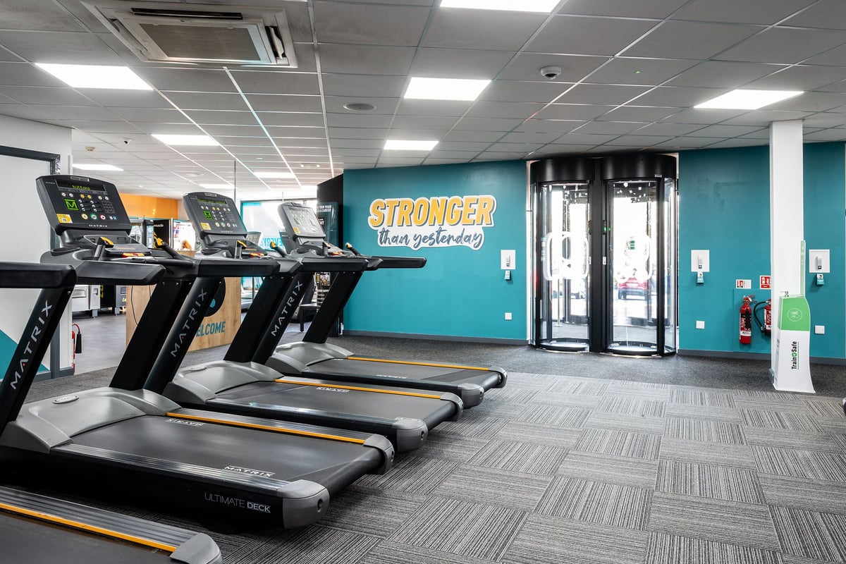PureGym Newtownabbey: Rows of treadmills, strength machines, and upright tanning booths in a modern gym interior.