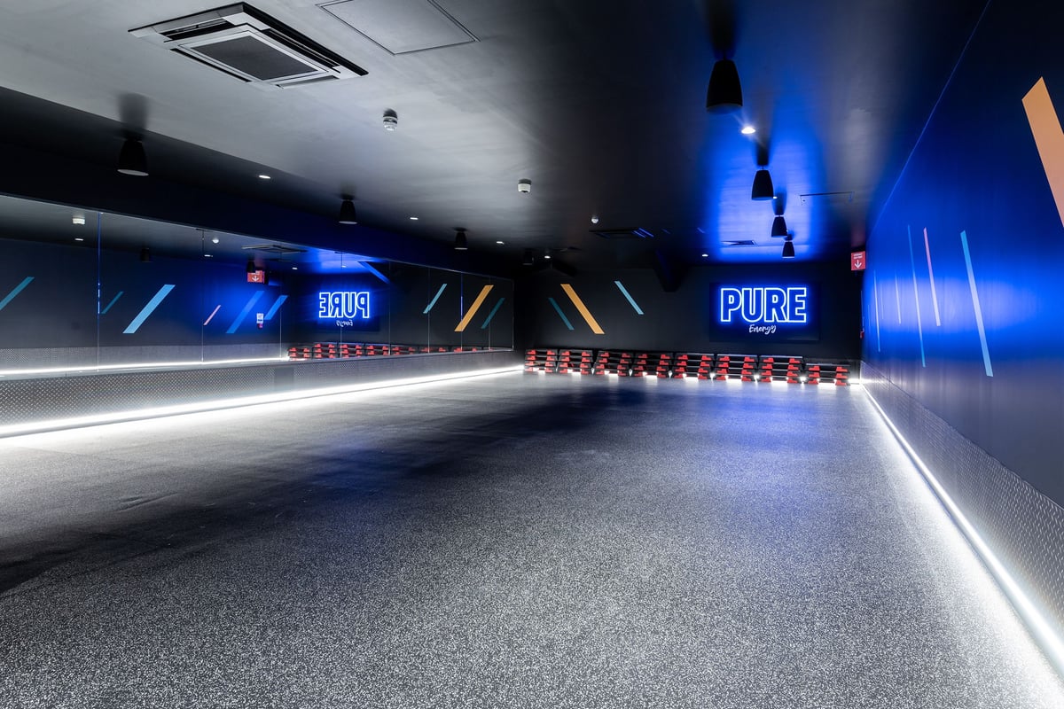 PureGym Bath Victoria Park: Fitness studio with mirrors, dark walls, and aerobic step platforms stacked against the far wall.