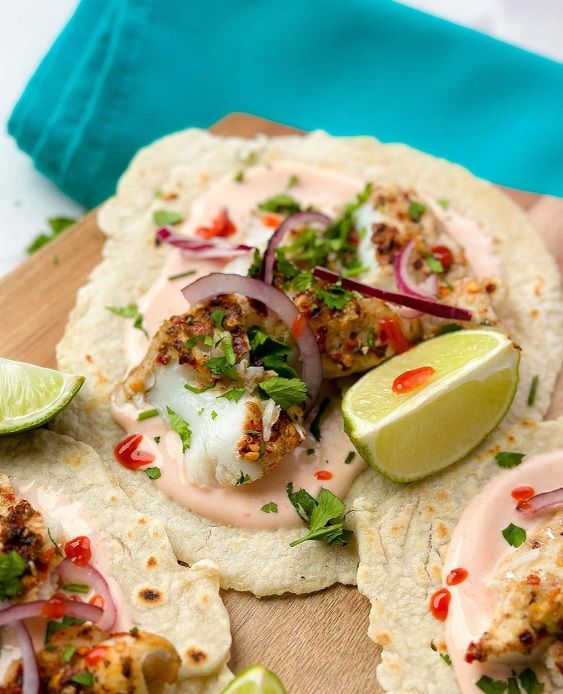 Fish tacos with sauce, red onion, herbs, and lime wedges on a wooden board