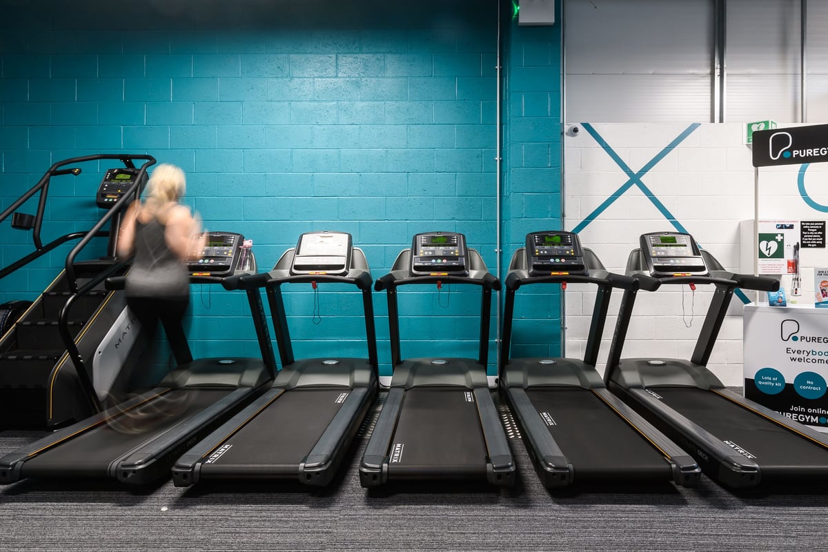 PureGym Buckingham: Rows of treadmills inside a teal-coloured gym with one person exercising.