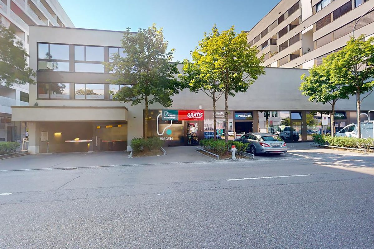 PureGym Schlieren: Exterior of building with shops and trees in front, entrance to underground parking visible.
