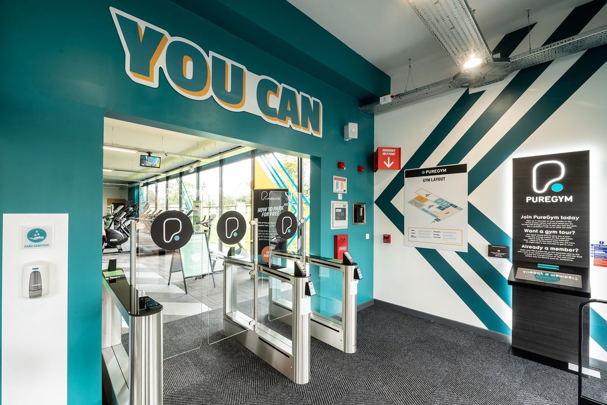 PureGym Witney: PureGym self-service kiosk area with turnstiles and promotional stands.
