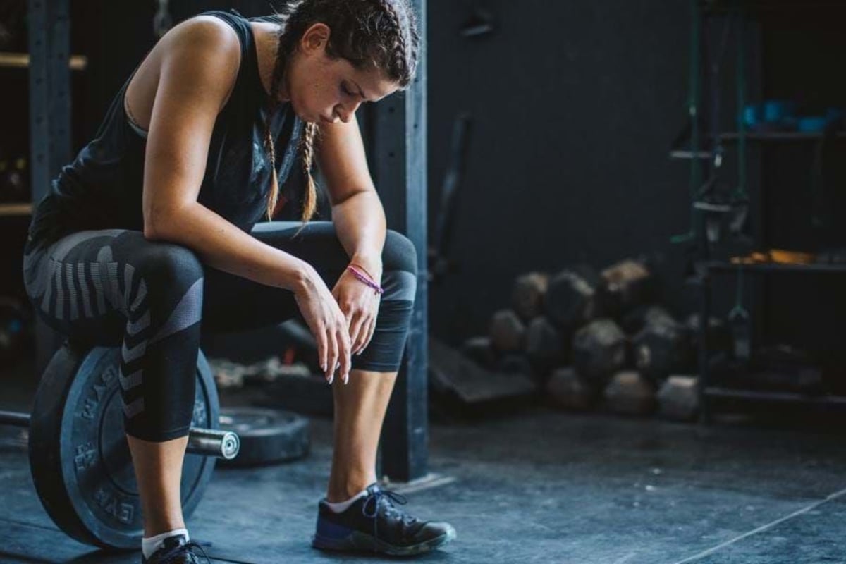 Woman in sportswear sitting on a barbell weight in a gym, looking down.