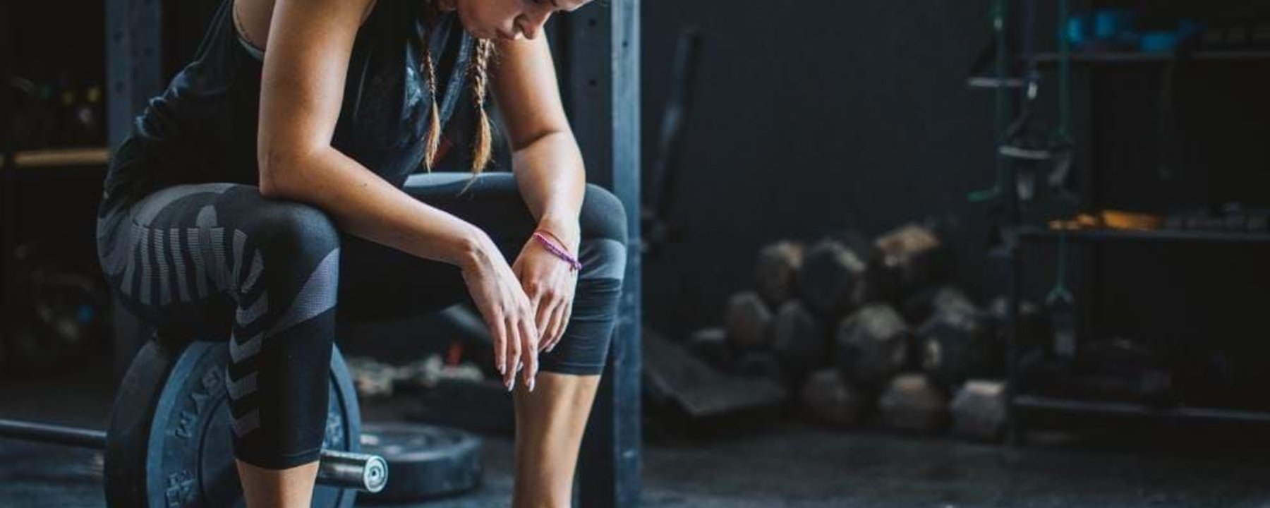 Woman in sportswear sitting on a barbell weight in a gym, looking down.