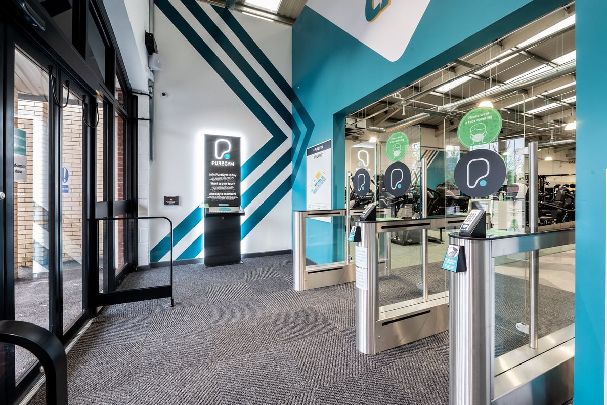PureGym Stockport North: Gym entrance area with turnstiles, kiosks displaying branding, and teal accented walls.