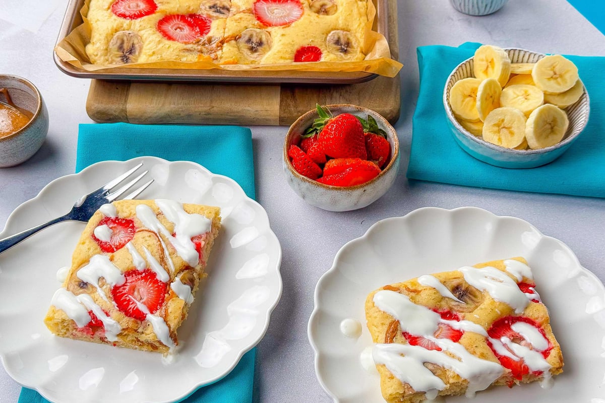Sliced strawberry and banana cake with white icing on two plates and bowls of fruit