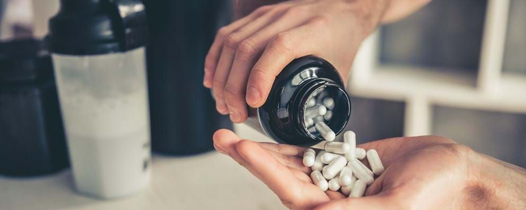 Hands pouring white capsules from a black bottle with shaker bottles in the background
