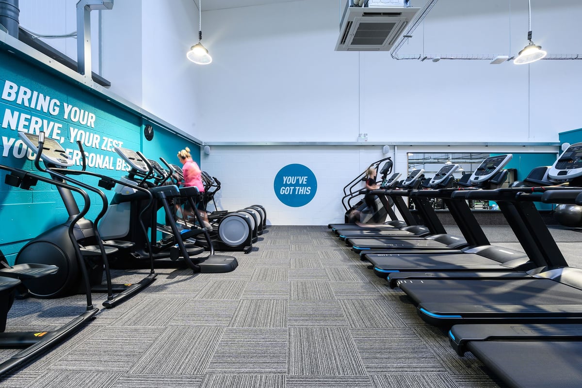 PureGym Saffron Walden: Rows of upright exercise bikes and treadmills in a gym studio with teal accents.