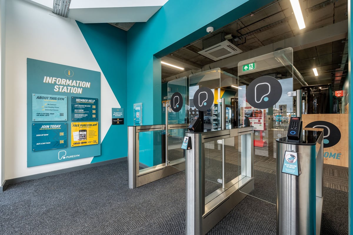 PureGym Brentwood: Information station, turnstiles, directional signage in turquoise and grey tones.