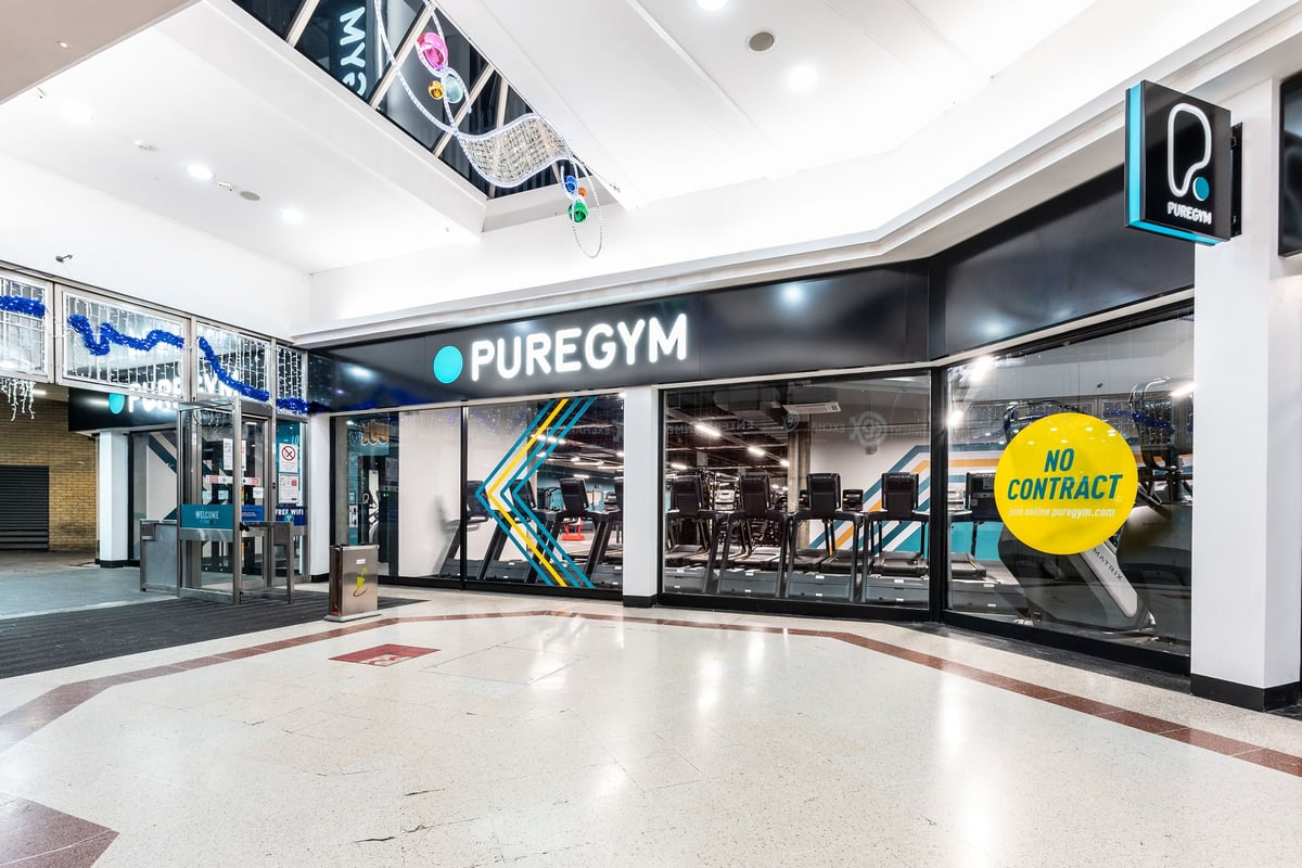 PureGym London Stratford: Glass front gym entrance, exercise equipment visible inside, yellow "No Contract" sign displayed.