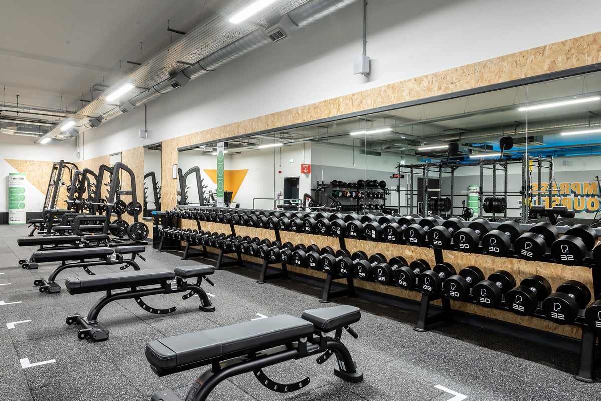 PureGym Witney: Rows of benches, racks stocked with free weights, rubberized flooring in a gym weight room.