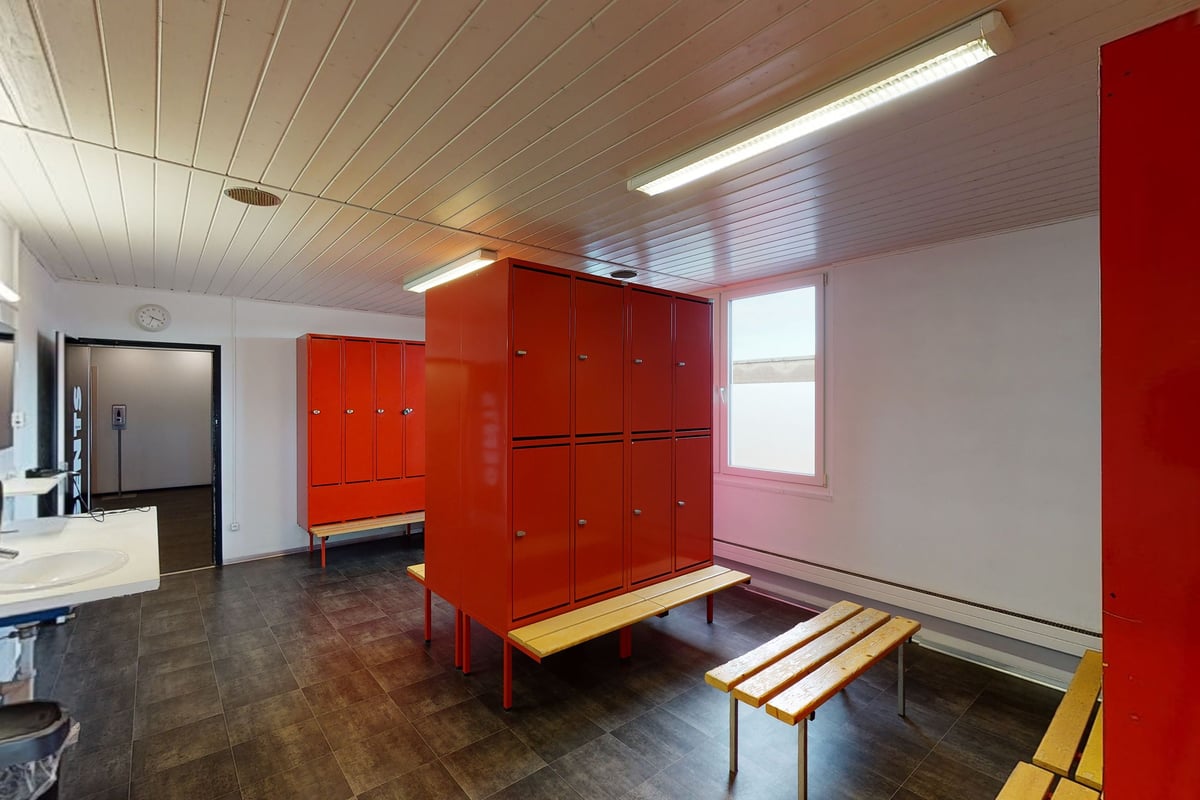 PureGym Rorschach: Vestiaire avec casiers rouges, bancs, télévision et plafond lambrissé avec éclairage fluorescent.