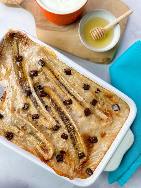 Baked banana dessert with chocolate chunks in a white dish beside honey and cream bowls