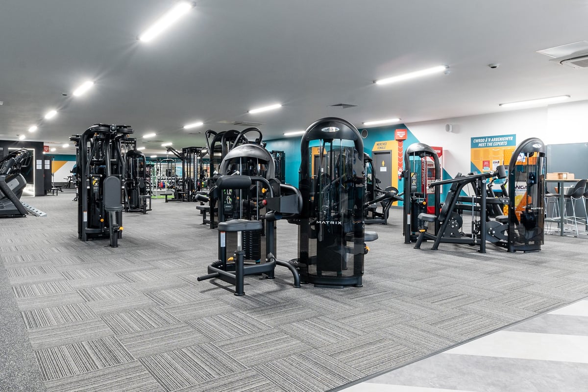 PureGym Bangor Wales: Rows of resistance machines, weight stacks, benches, and rubberized flooring in a gym setting.