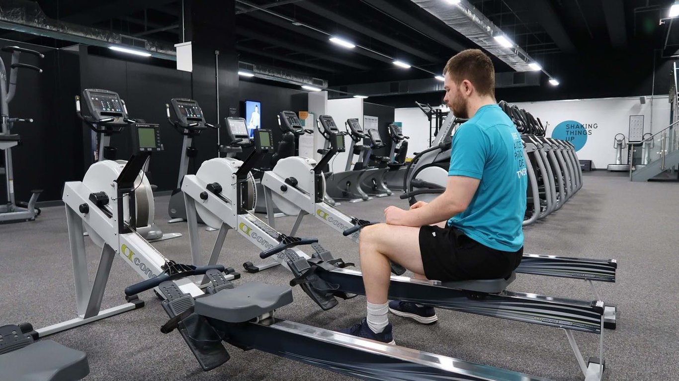 Man in blue shirt sitting on rowing machine in gym with exercise bikes in background