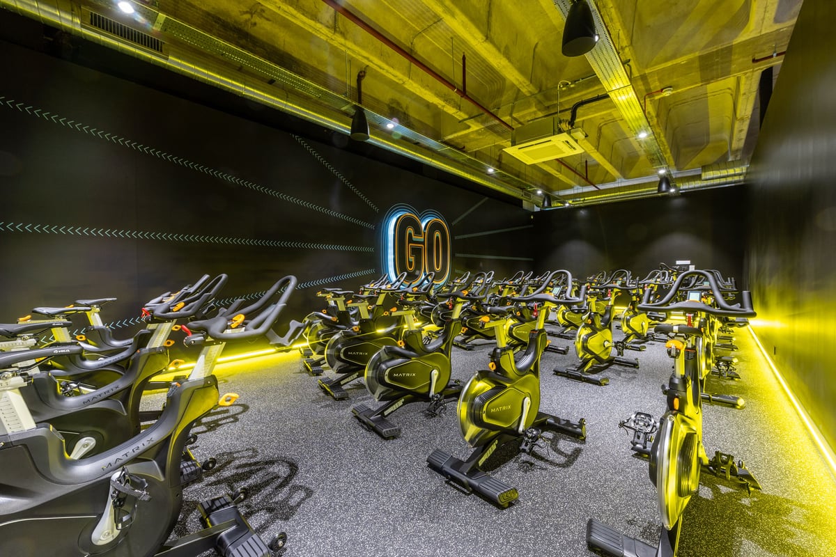 PureGym London Ealing Broadway: Rows of yellow spin bikes in a dimly lit studio with neon 'Go' sign.