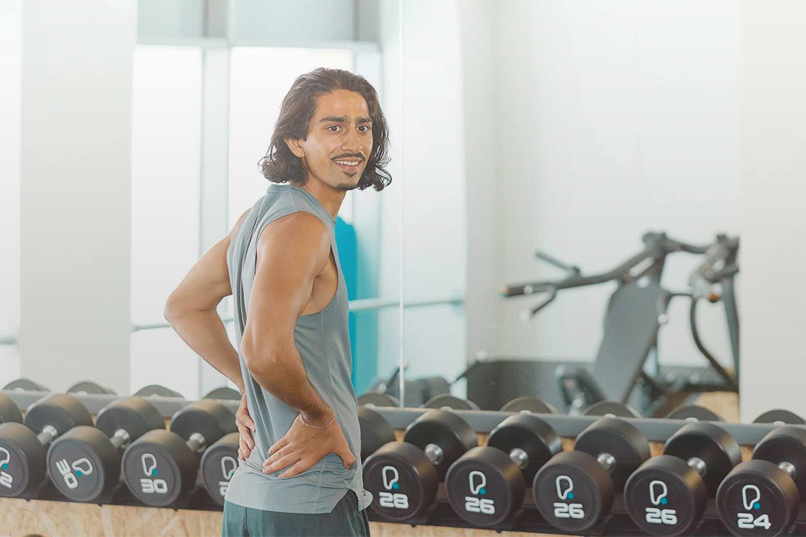 Mann mit langen dunklen Haaren lächelt vor einem Hantelregal im PureGym Schweiz
