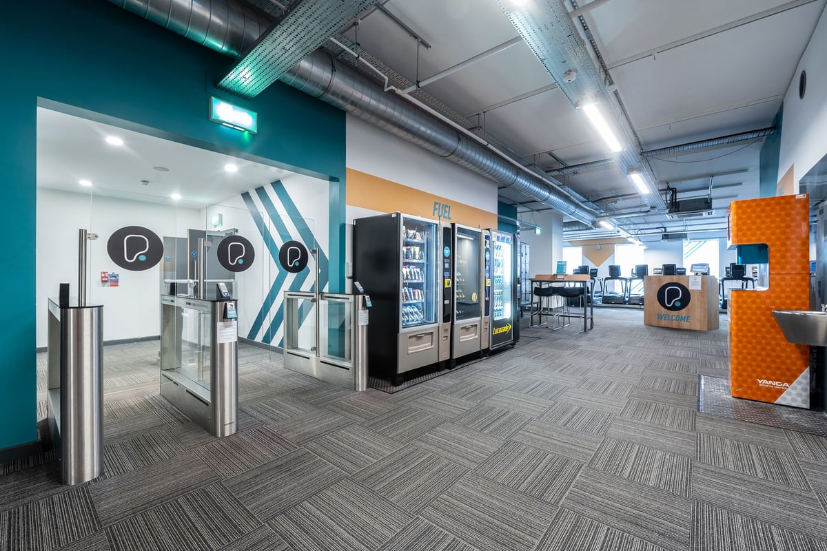 PureGym Stevenage: Entry area with turnstiles, vending machines, and seating in a modern fitness centre.