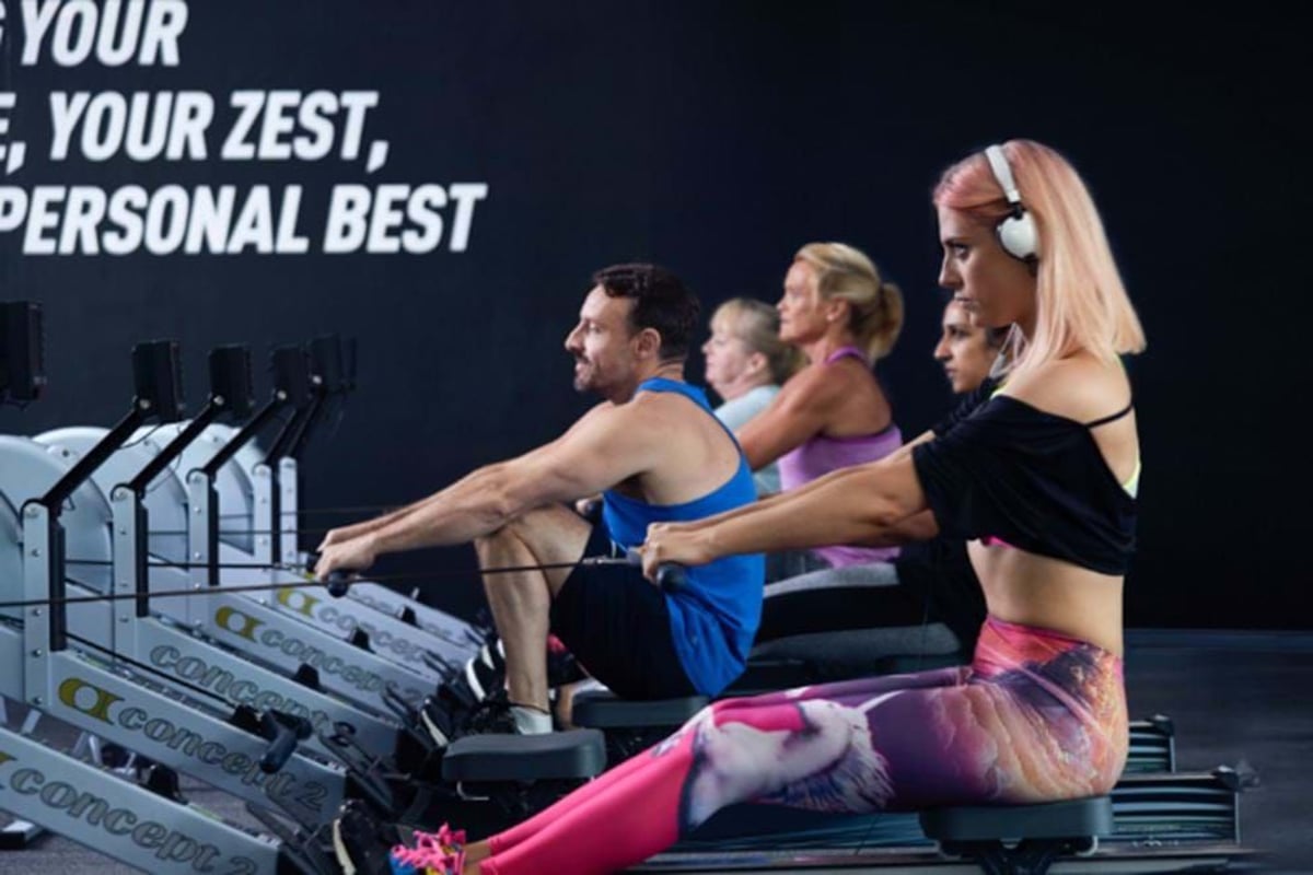 Five people exercising on rowing machines in a gym with motivational text on the wall