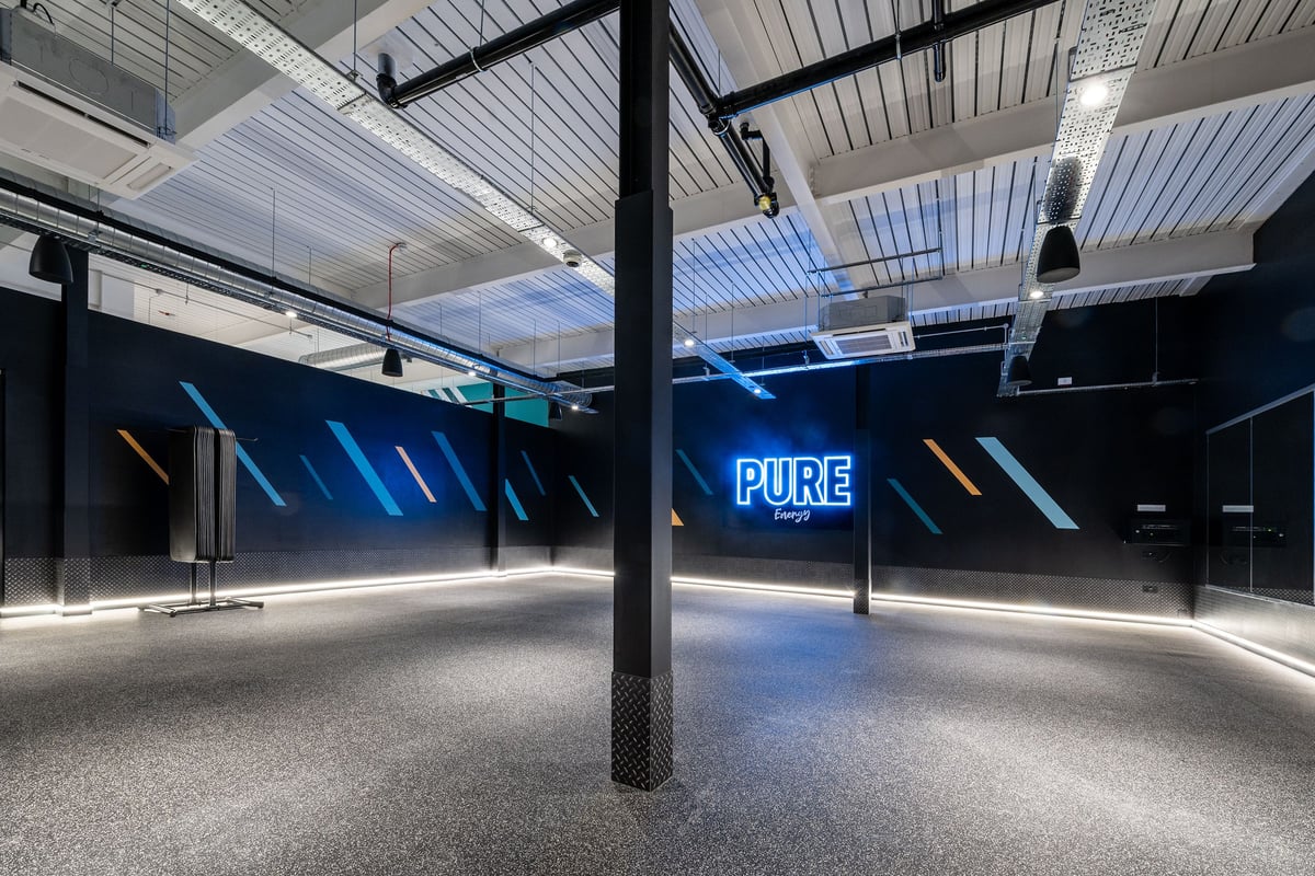 PureGym London Colney: Dimly lit studio interior with blue neon signage, steel beams, and rubberized flooring.