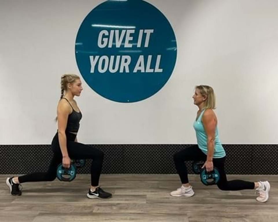 Two women doing lunges with weights in front of a wall sign saying Give it your all