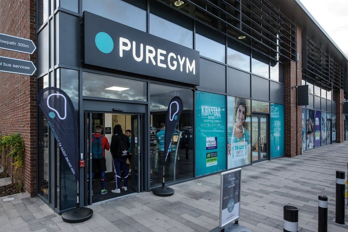 PureGym Leeds Kirkstall Bridge: Puregym storefront with glass windows, signage, and exterior seating and advertising boards.