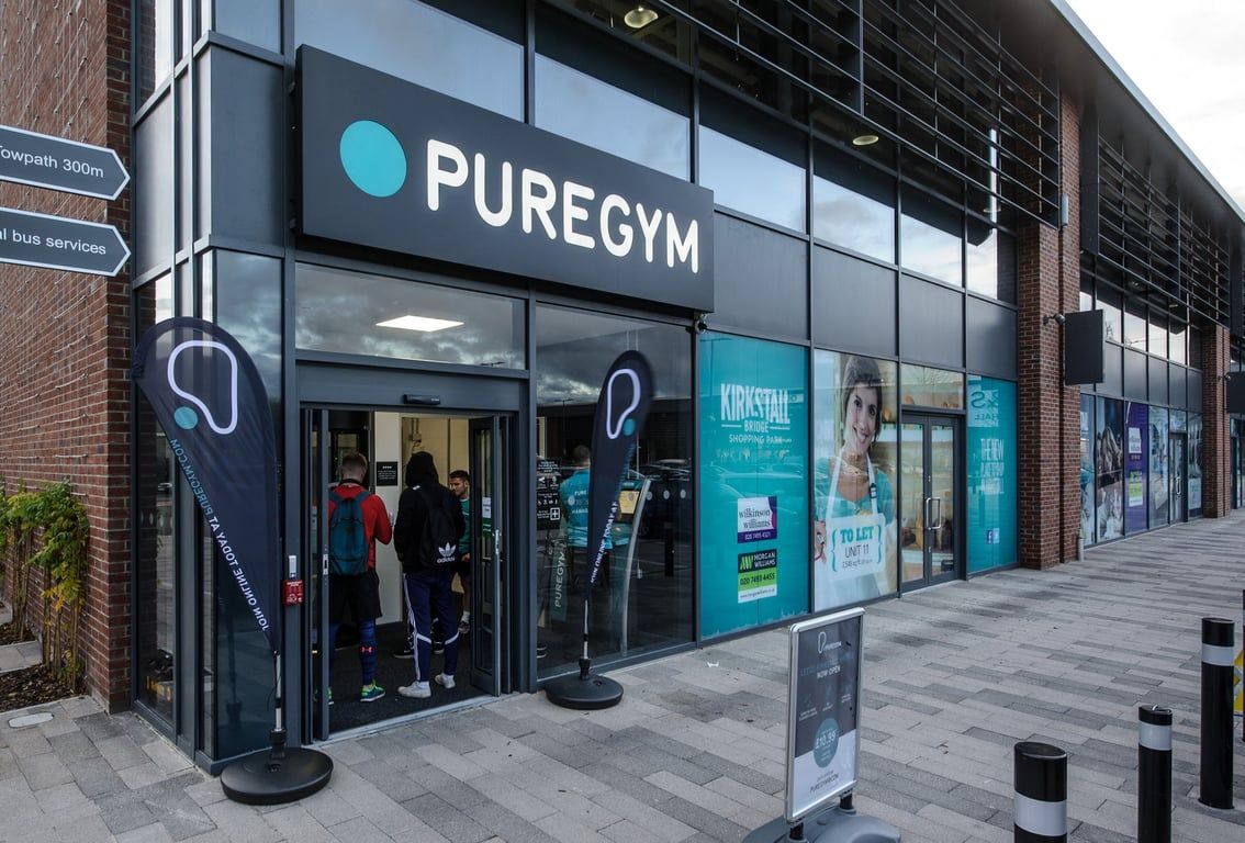 PureGym Leeds Kirkstall Bridge: Puregym storefront with glass windows, signage, and exterior seating and advertising boards.