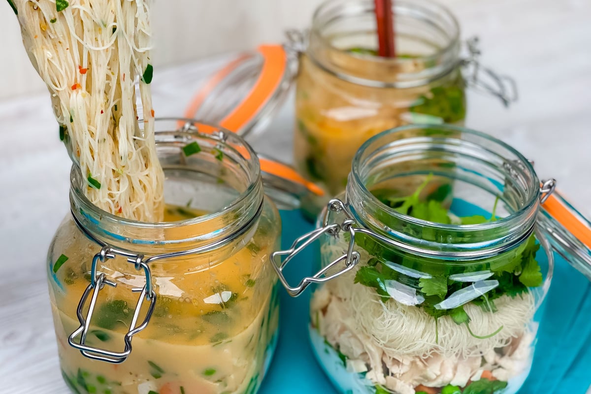 Rice noodles being lifted from glass jar filled with broth and vegetables