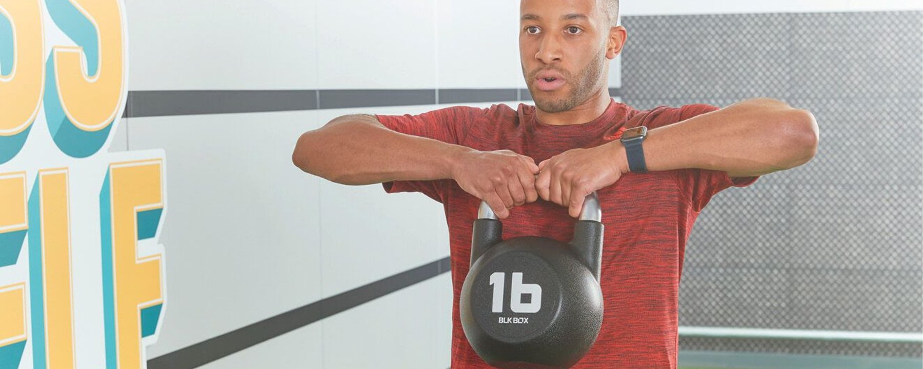 Man in red shirt lifting a 16-pound kettlebell in a gym with motivational wall art