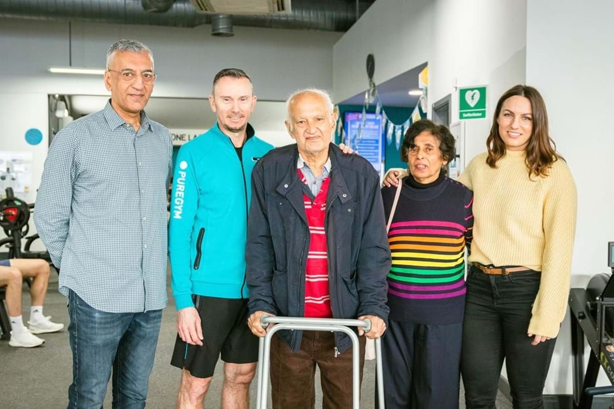 Five people standing together in a gym, one elderly man using a walker.