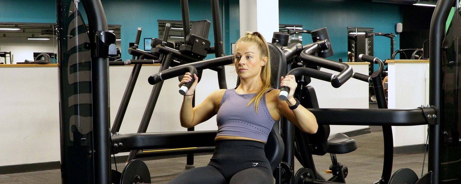 Woman in purple sports top using chest press machine in gym with turquoise walls