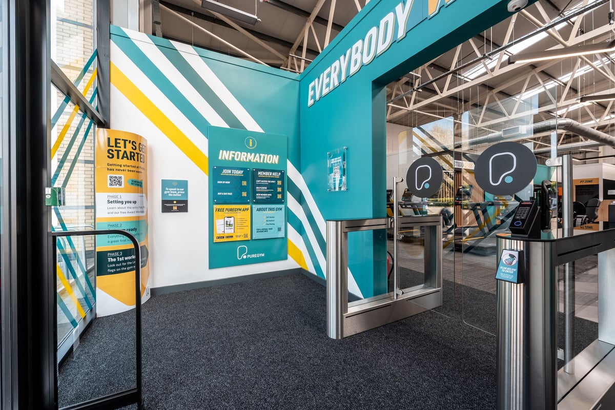 PureGym Bridgend: Teal-colored information kiosk, signage promoting gym memberships, and entry turnstiles.