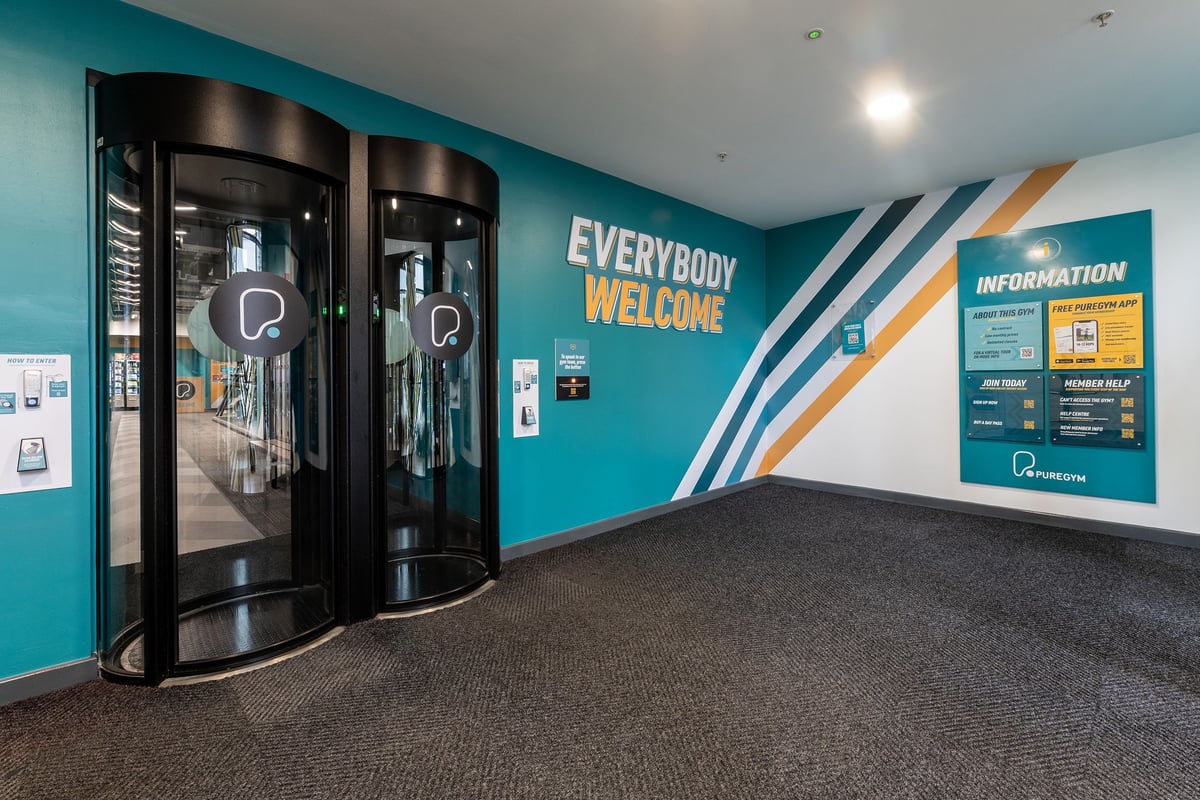 PureGym Ipswich Buttermarket: Glass entrance doors, teal walls with "Everybody Welcome!" text, information kiosk.