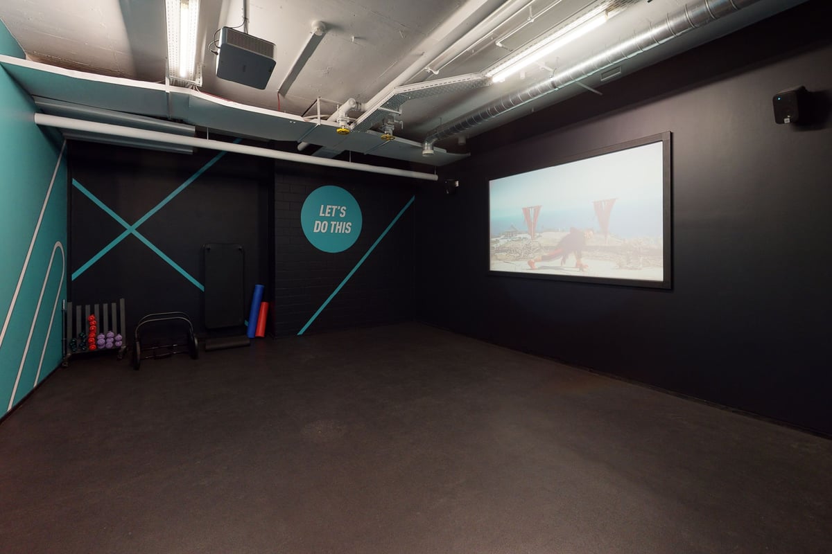 PureGym Bern Bümpliz: Studio vide avec écran de projection, touches bleu sarcelle, murs noirs et sol caoutchouté.
