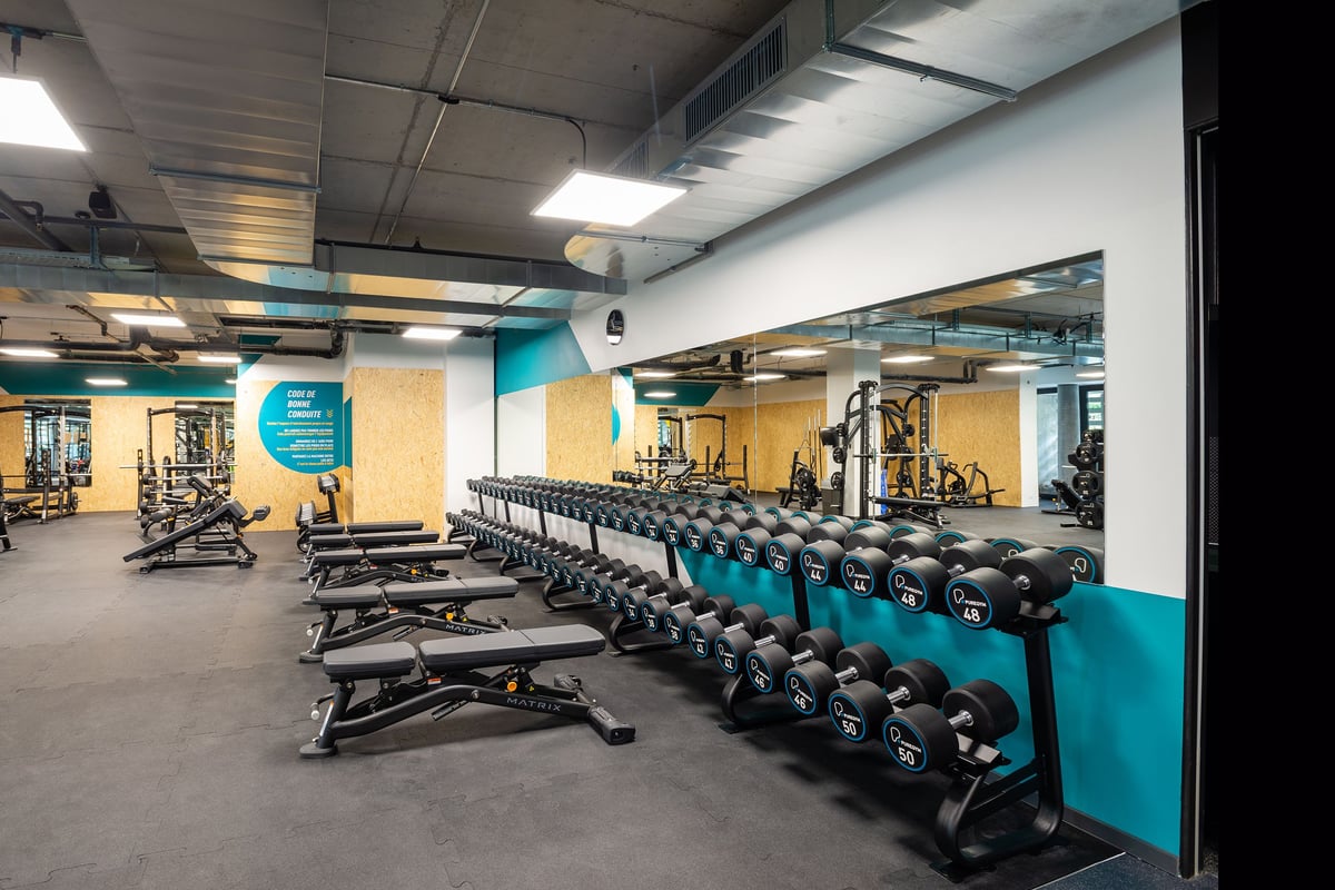 PureGym Genf Le Bouchet: Rows of benches, dumbbells in teal racks, rubberized flooring, and weightlifting equipment.
