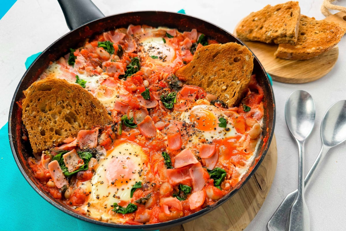 Shakshuka with eggs, spinach, ham, and toasted bread in a frying pan with spoons nearby